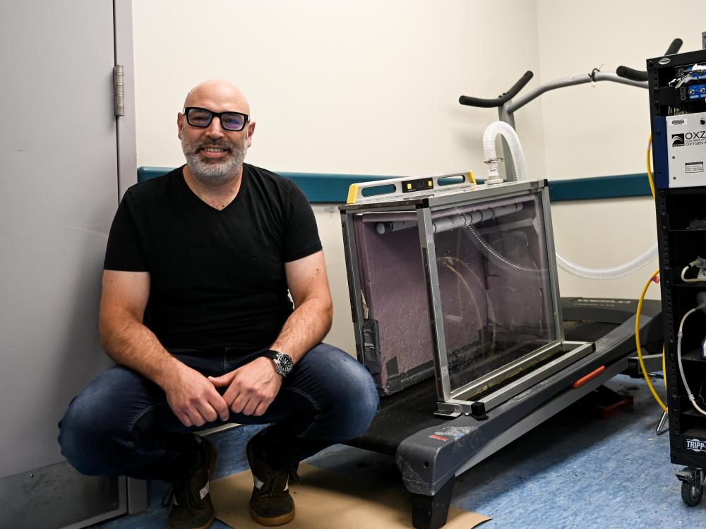 Jonas Rubenson crouches beside a treadmill. On the treadmill is the metabolic chamber, a Lucite box with tubes for airflow.