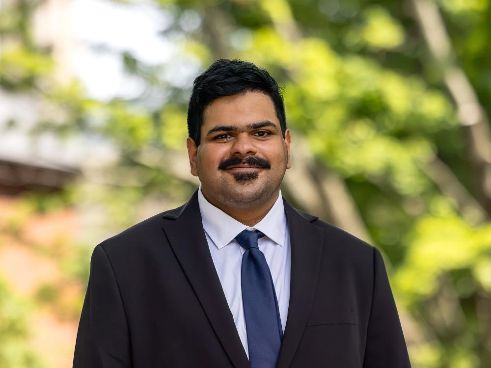 Hamad Hussain wears a black suit jacket, white shirt, and navy blue tie while standing in front of a tree.