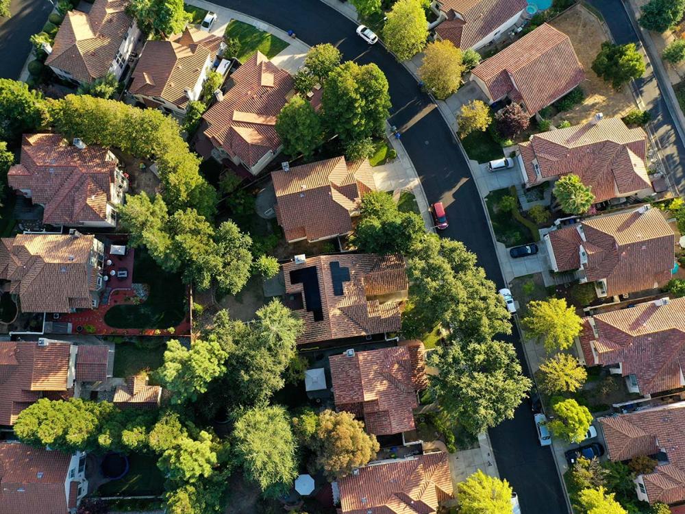 Aerial view of a suburban neighborhood with red-tiled roofs, surrounded by green trees and neatly paved streets, showing several homes and yards.