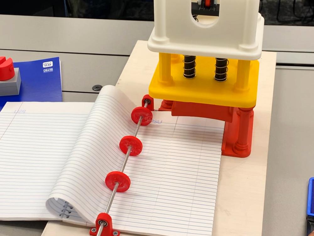 Notebook beside a colorful 3D-printed device with red rods and discs on a desk.