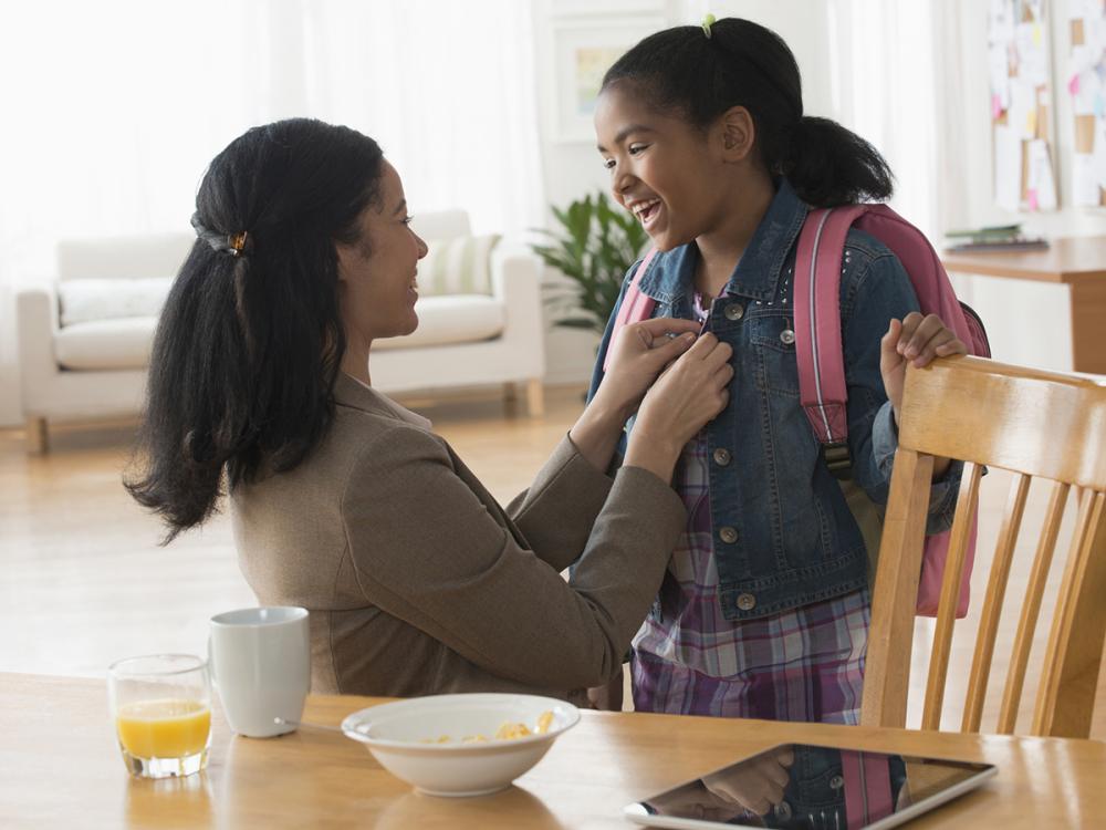Mother buttons daughter’s jacket as she gets ready for school. Daughter is wearing a backpack. A table with breakfast dishes is in front of them.