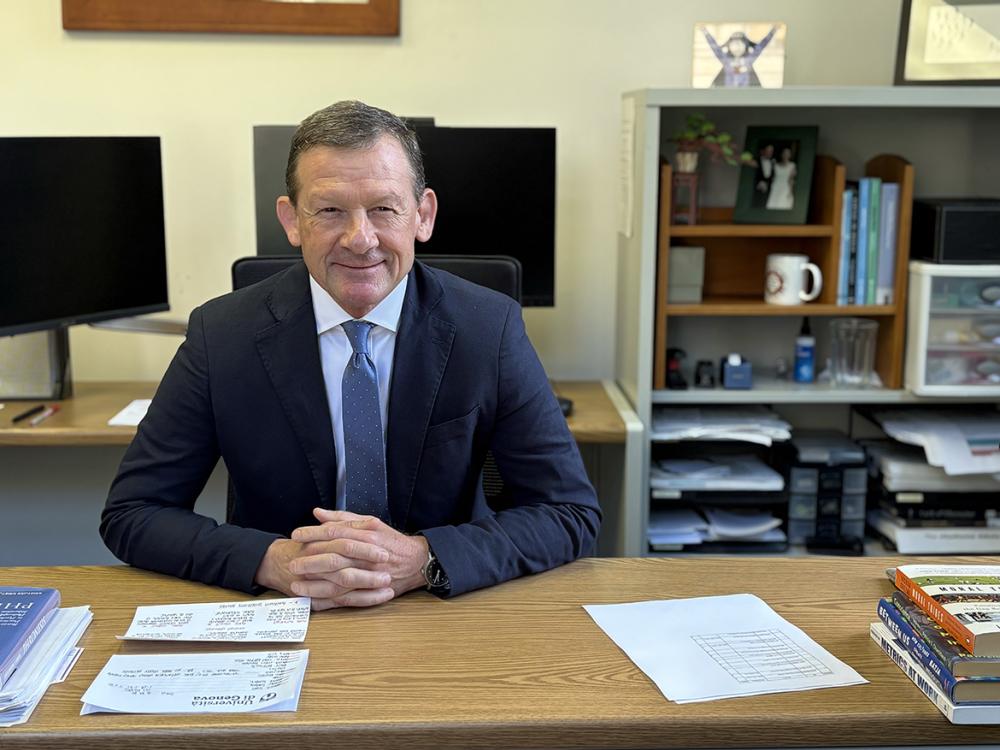 A man in a suit and tie sits at a desk and smiles at the camera.
