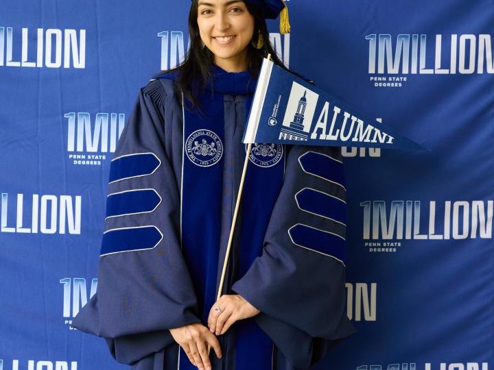 A student wearing regalia poses in front of a backdrop that reads "1MILLION" while holding a pennant that reads "Alumni"