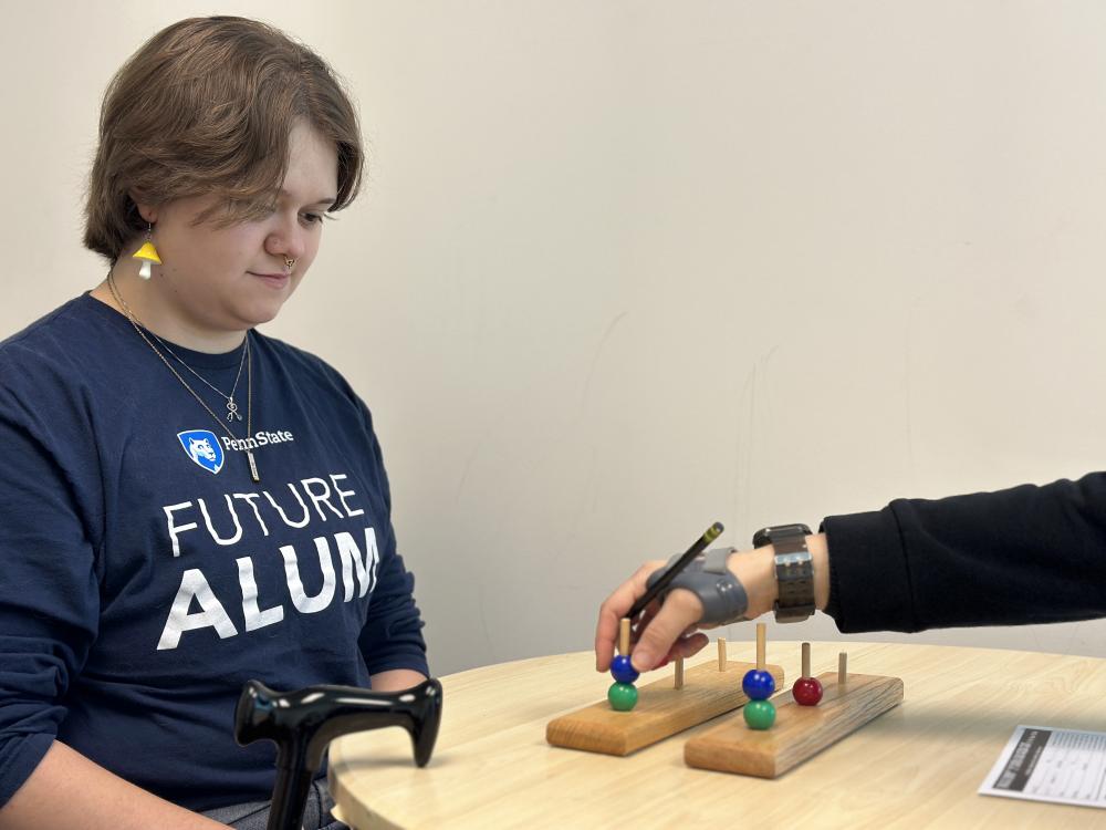 A student sits at a table watching as a hand (the person is out of the shot) moves bright wooden balls on a peg board