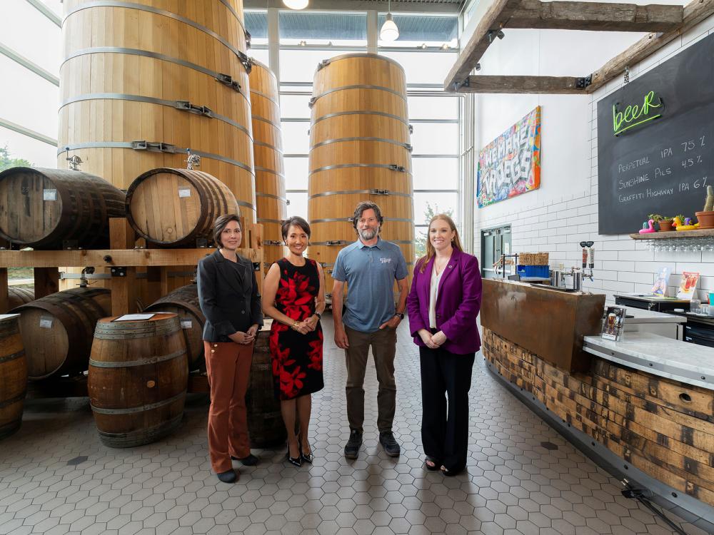 Four people stand in a large room with wooden barrel kegs stacked on one side