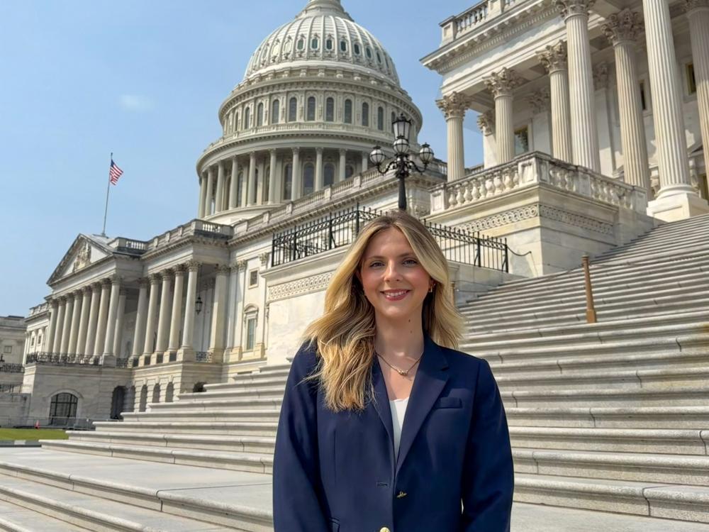 Alivia Reid-Pitta stands in front of the United States Capitol wearing a navy blazer and white top. 