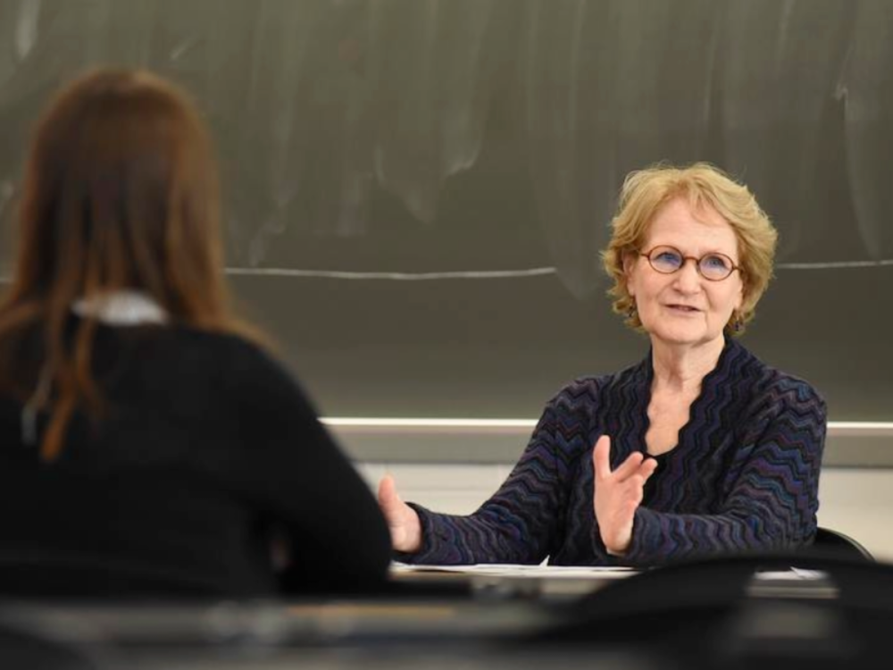 Barbara Rolls is pictured sitting at a desk at the front of a classroom, we can see the back of a student who is seated facing the front of the room.