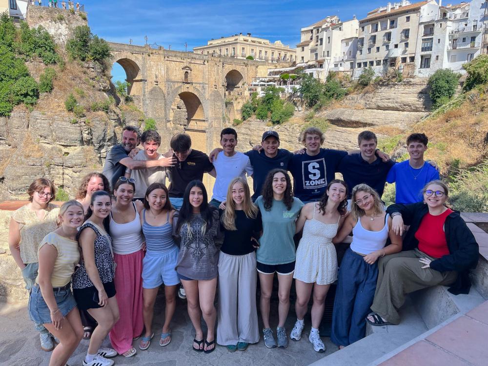 A group of Penn State students standing together, posed against a backdrop of a picturesque, historical hillside with colorful buildings and a stone bridge.