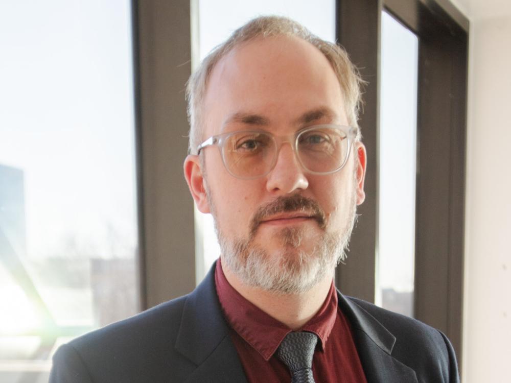 Brandon Sherman wearing a dark suit, burgundy shirt, and dark tie. 