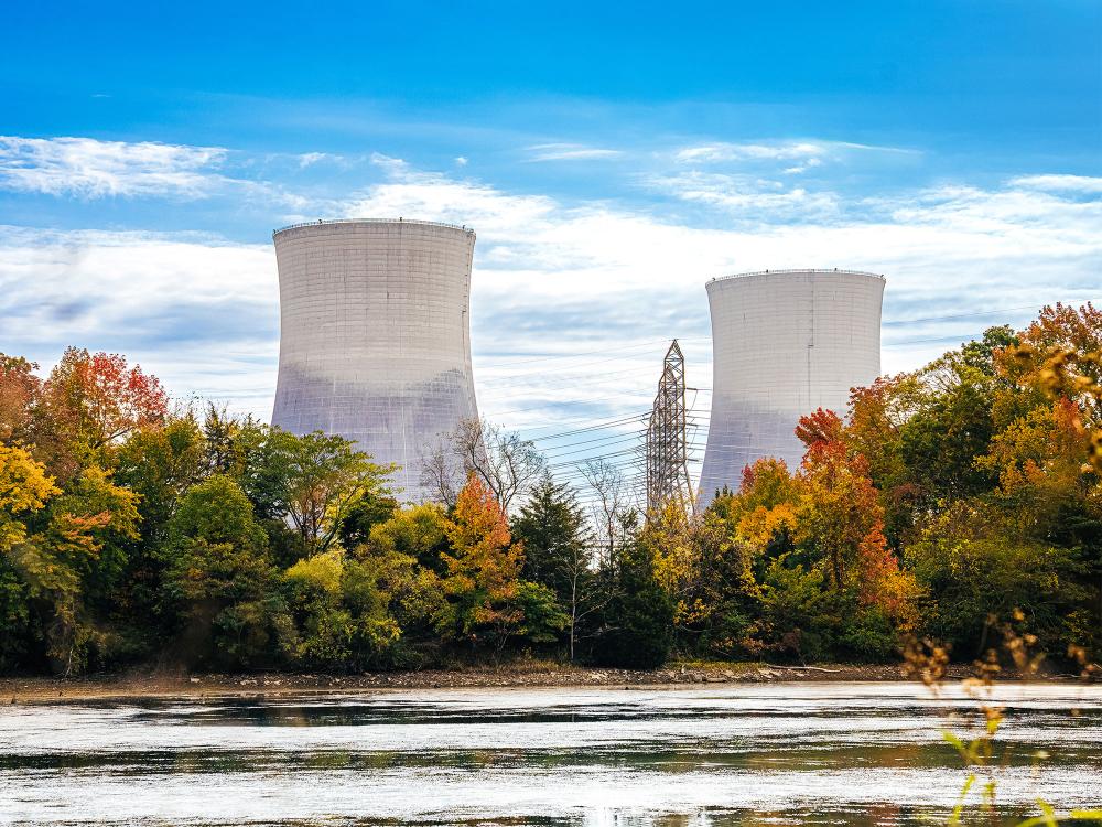 A view over a lake shows two large cooling towers of a nuclear power plant overlooking some trees on a sunny day
