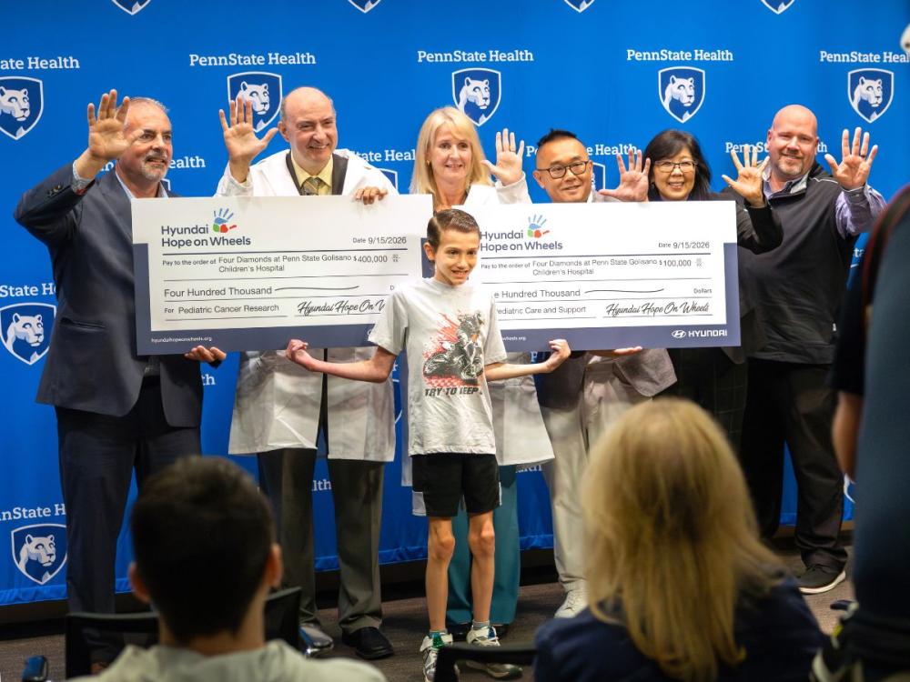 Group of Penn State Health physicians and staff hold oversized Hyundai Hope on Wheels checks totaling $500,000 for pediatric cancer research and care, with a child standing in front of the group.