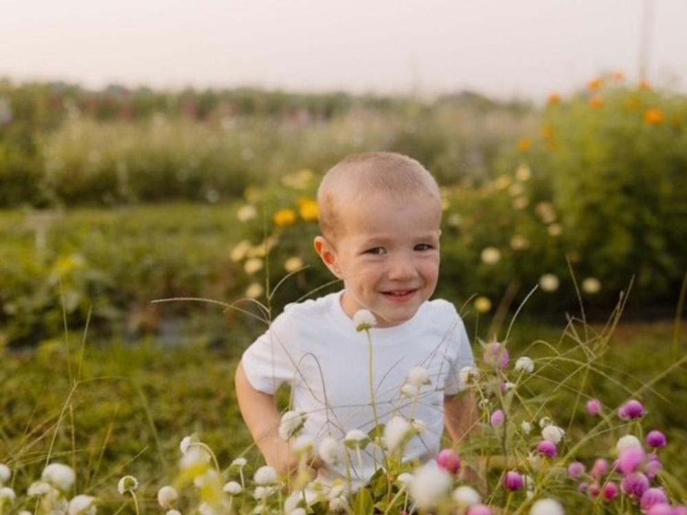 A young child wearing a white T-shirt sits among blooming wildflowers in an outdoor garden or flower field.
