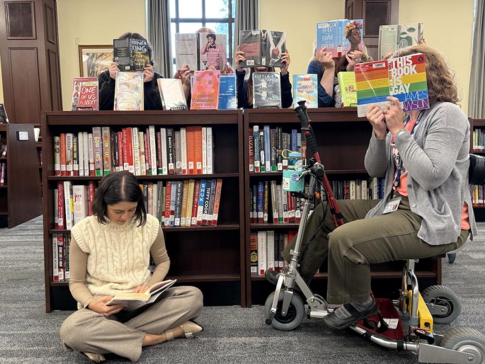 individuals sitting and standing with banned books open in front of their faces, around a bookshelf