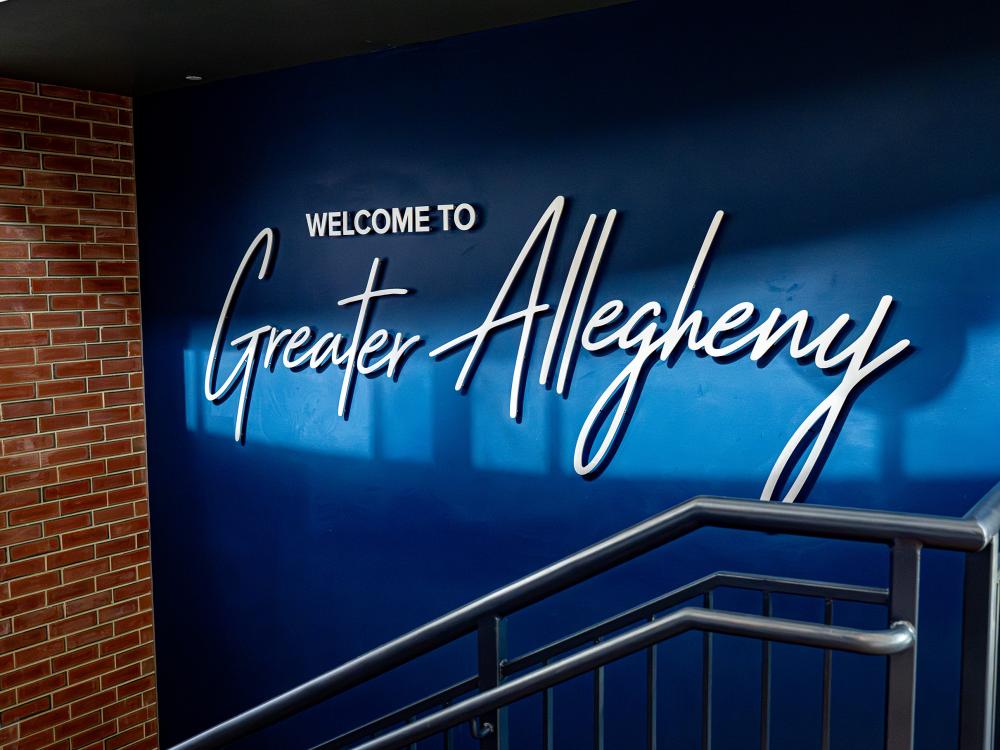 Interior stairwell featuring a blue wall with large white lettering that reads “Welcome to Greater Allegheny.” A gray metal handrail runs along the stairs in the foreground, with a red brick wall visible on the left.