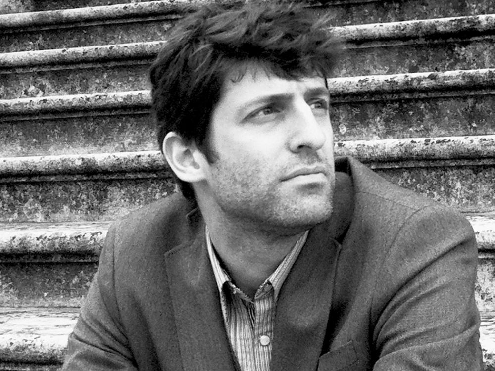 Black and white portrait of a man in a suit jacket, seated on weathered stone steps and gazing off to the side.