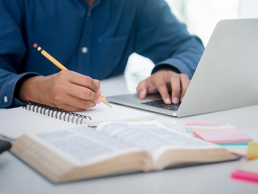 Person writes in a notebook while using a laptop, with an open book and sticky notes on the desk.