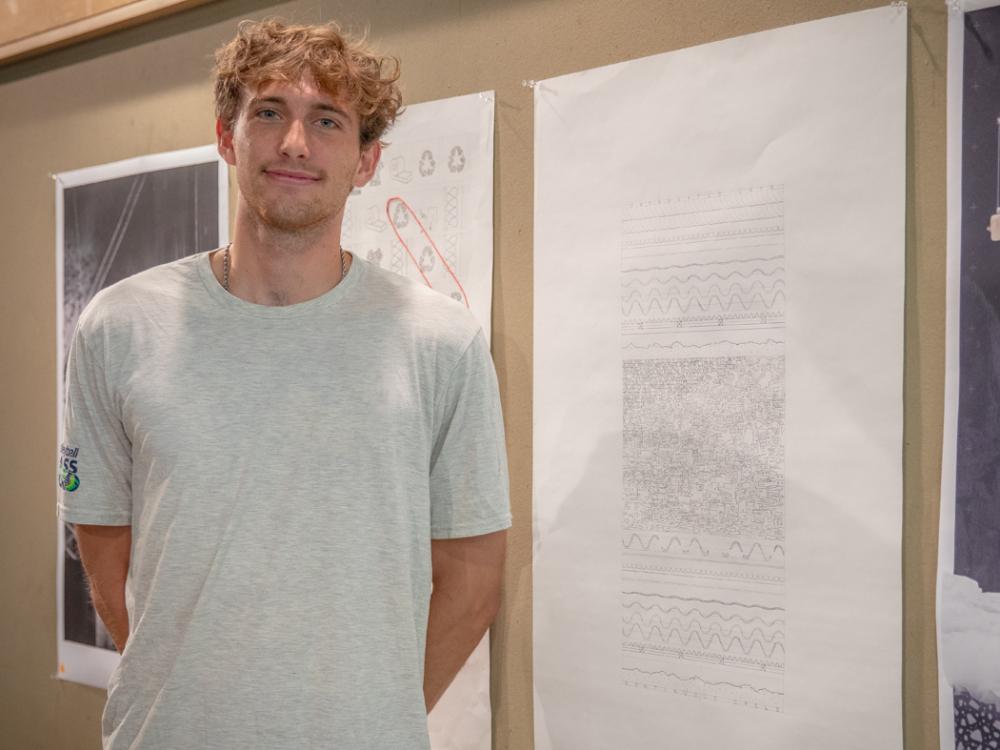 Young man standing beside a large abstract drawing displayed on a gallery wall, with additional artwork visible in the background.