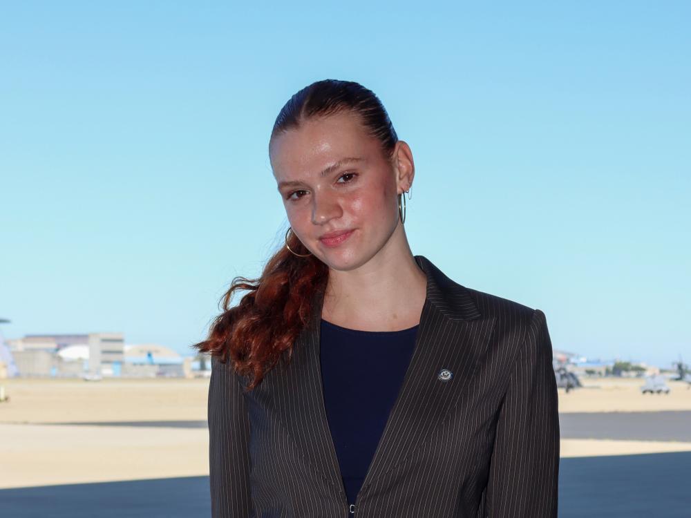 Odessa Dando wearing a dark pinstripe suit standing on a clear, open area with a distant, partly visible federal airfield in the background.