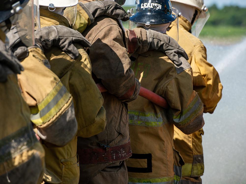 Firefighters in protective gear work together near a road, carrying a hose.