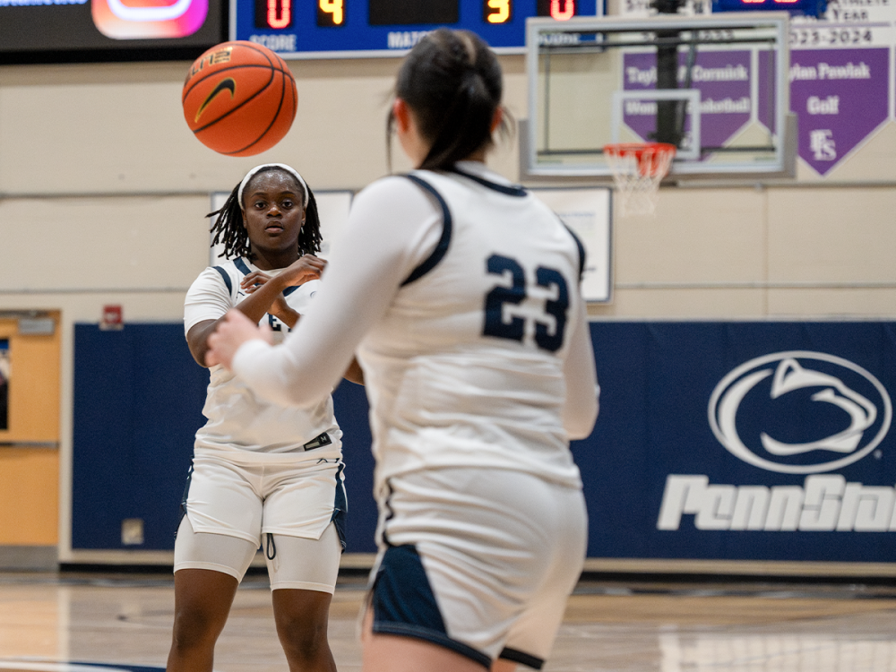 Two Penn State Fayette basketball players in white uniforms passed a basketball on the court. The player closest to the camera wore No. 23; a hoop and Penn State signage were visible behind them.