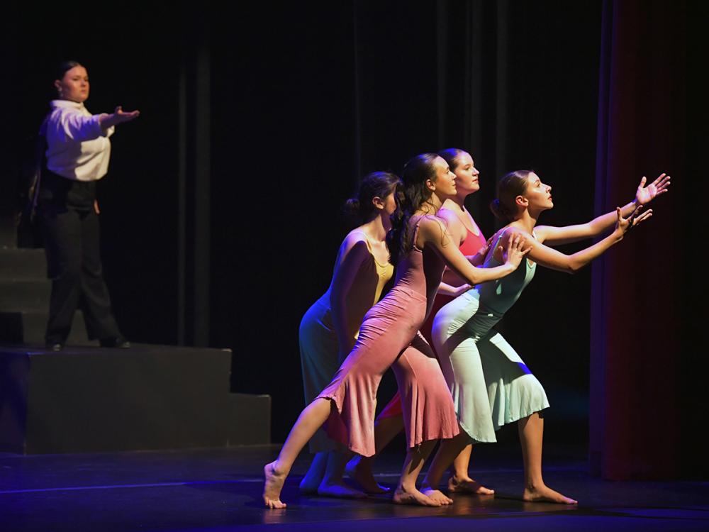 Five dancers perform on a dimly lit stage, reaching forward in colorful costumes.