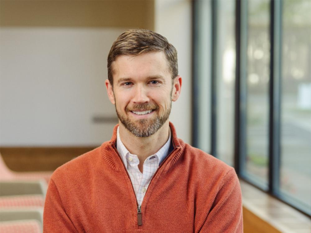 A man with light brown hair and beard wearing a red-orange quarter zip sweater