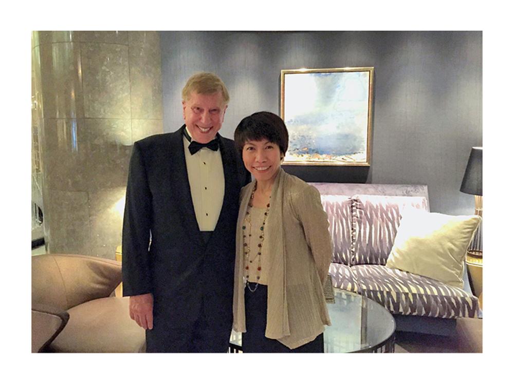 A man with light colored hair wearing a tuxedo and smiling while standing next to a petite woman in a tan dress smiling