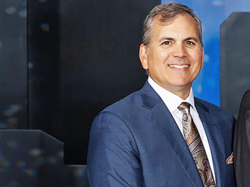 A photo of a smiling Joe Abraham pictured from chest up in a blue suit and multi-colored tie