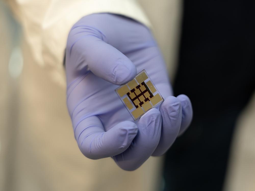 A gloved hand displays a small, square object that resembles a tiny solar cell.