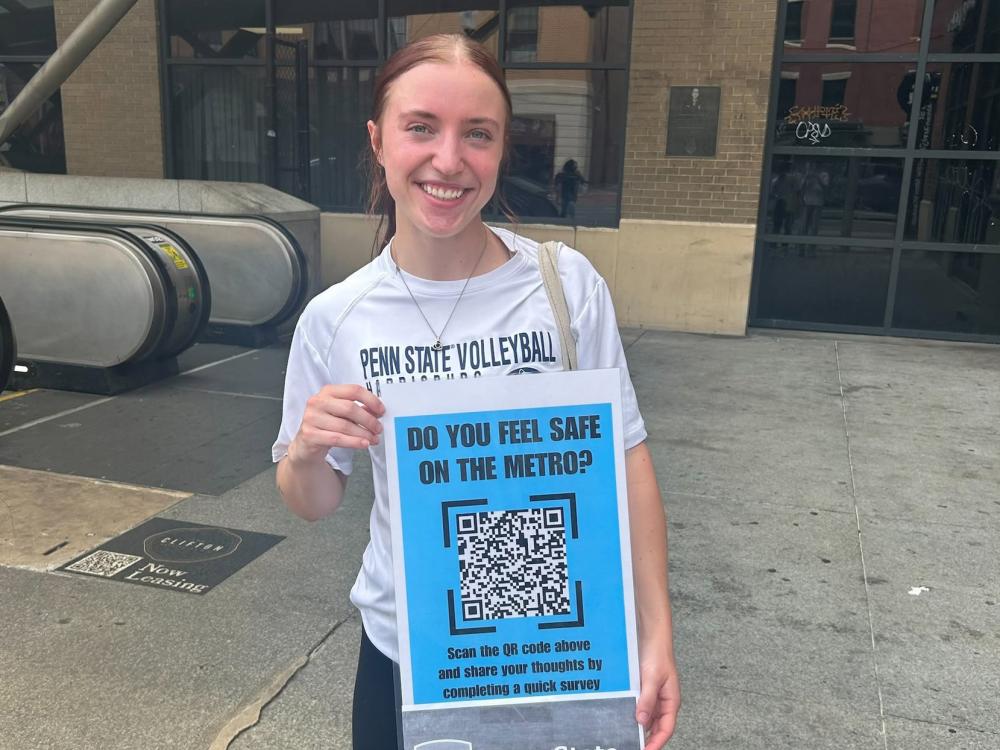 Kate Bukowski stands outside a metro station in DC with a poster inviting riders to take her survey