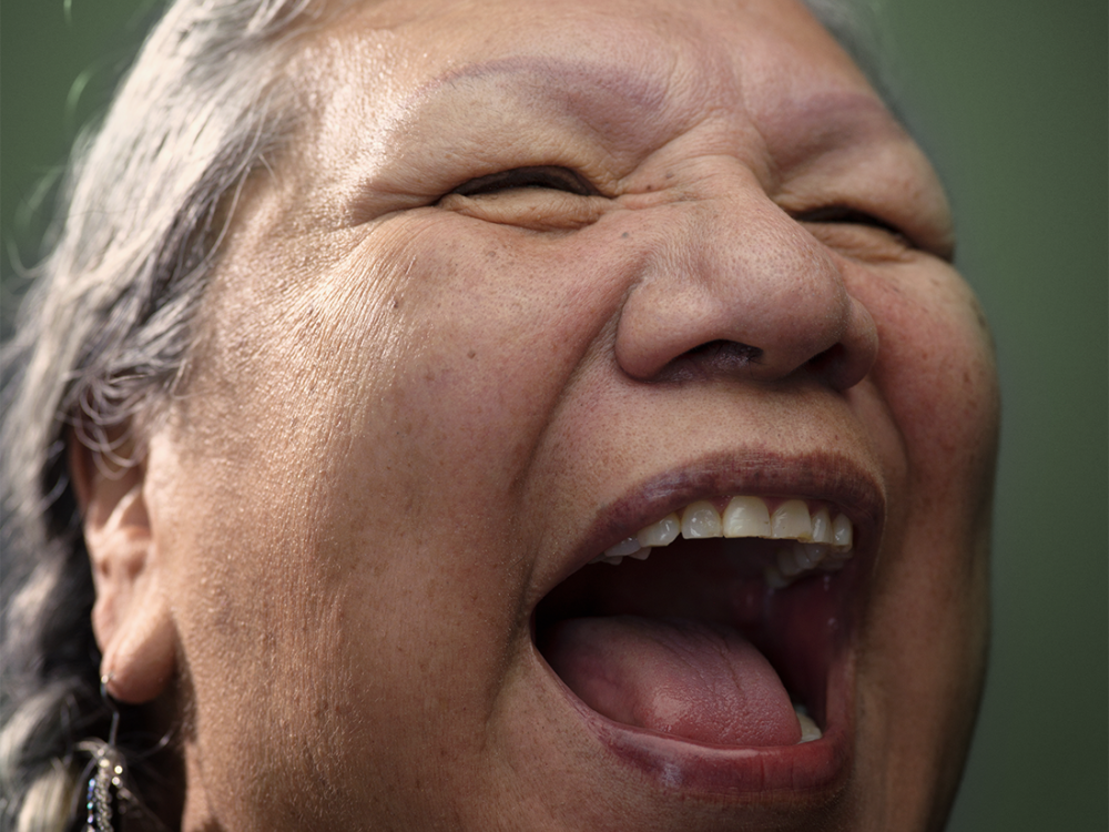 Photo portrait of Lakota Harden, Native community leader and elder, laughing broadly, open-mouthed. Her face, which makes up most of the image, is framed by gray hair in braids, dangling blue earrings, and a dark green background.