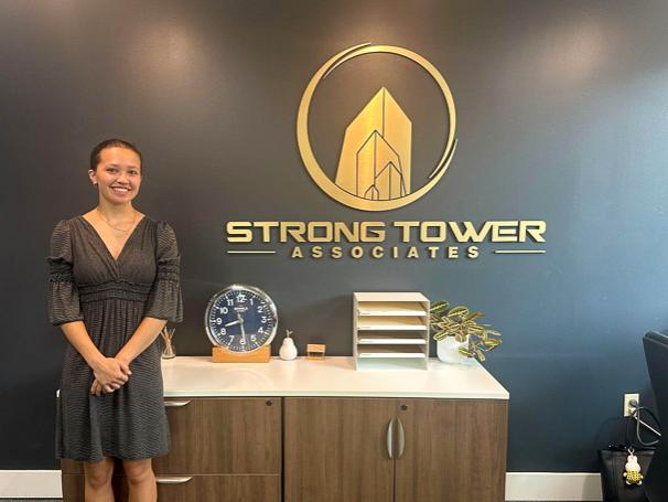 Sonia Lloyd wears a black dress and stands in front of the Strong Tower Associates sign. 