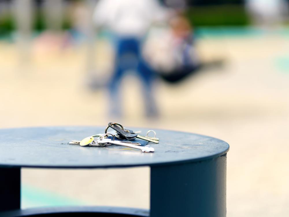 A set of keys sits on top of a trash can at an outdoor playground. Figures in soft focus play in the background.