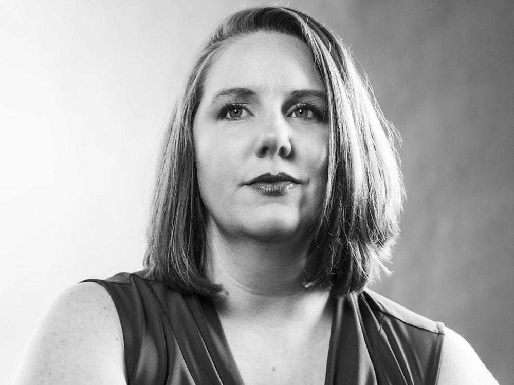 Black-and-white head-and-shoulders portrait of a person with shoulder-length hair, facing slightly to the side, wearing a sleeveless top against a softly blurred background.