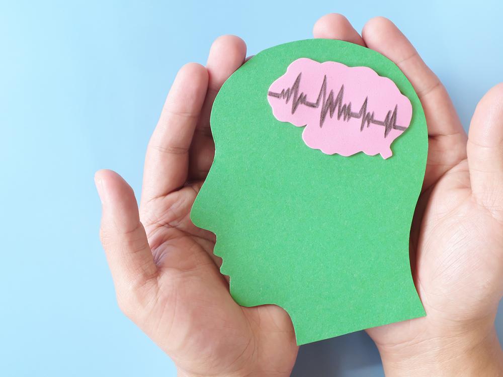 Hands hold a green head silhouette with a pink brain and heartbeat line.