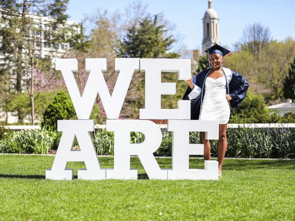 Nia Smith poses in graduation attire next to white block letter spelling “We Are” with the Old Main bell tower in the distance.