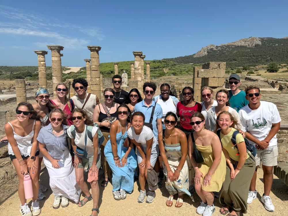 A group of people gather outdoors in front of ancient stone columns, surrounded by greenery, hills and a mountain under a clear blue sky.