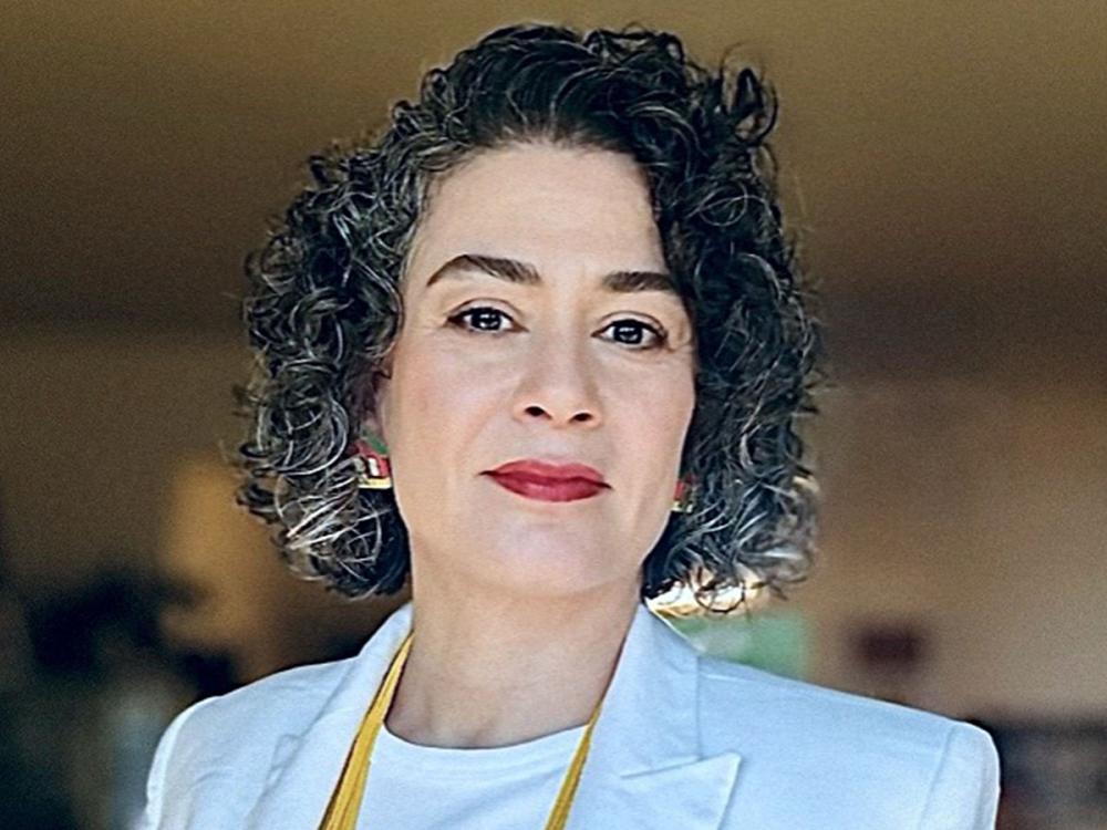 Portrait of Roberta Mendonça De Carvalho wearing a light blazer, white shirt, and yellow lanyard, facing the camera.