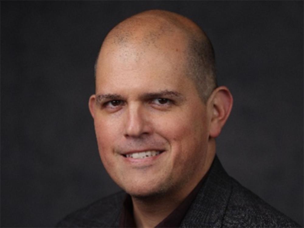 A bald man in a dark shirt in front of a dark background
