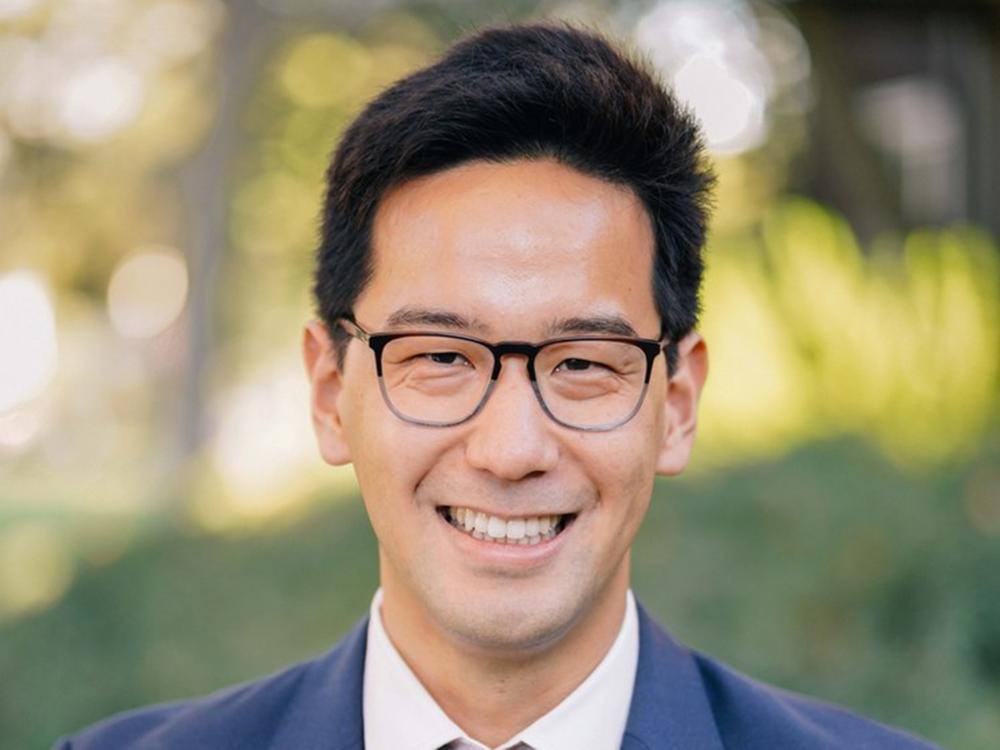 An asian man with dark hair and glasses smiling. Blurred greenery in the background