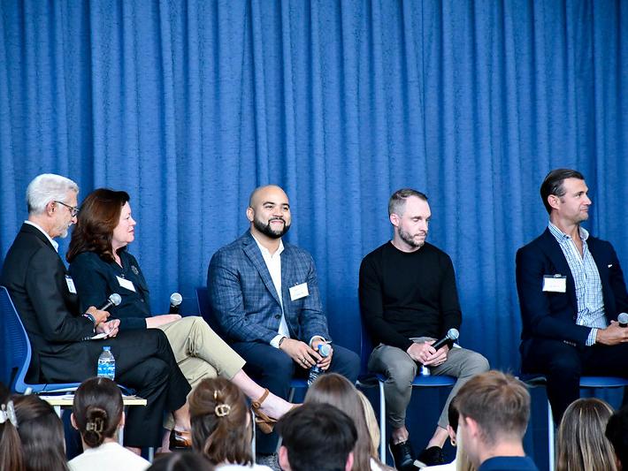 Five panelists, four men and a woman, sit onstage in front of a blue curtain while an audience watches.