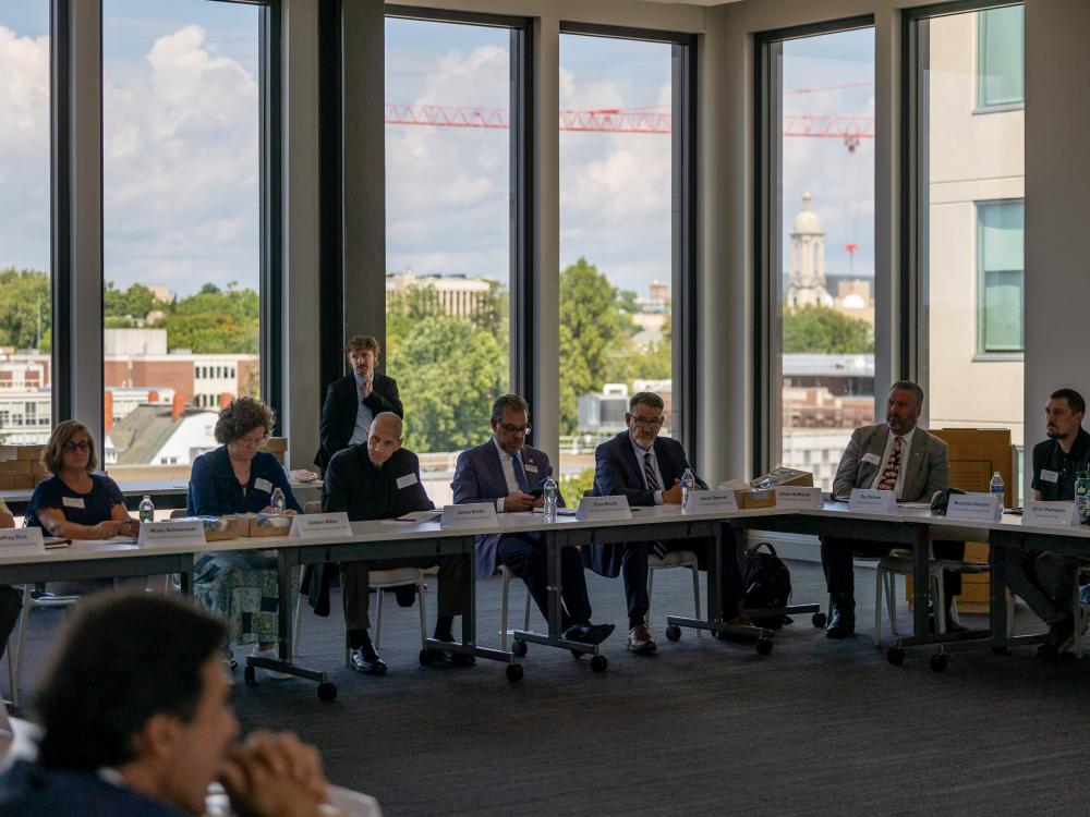 A row of people in business attire sit in front of a large window with a view of the Penn State Campus