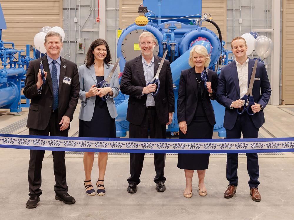 Leadership from industry, government and academia pose in front of a giant mechanical compressor, holding big scissors to cut a START+ branded ribbon