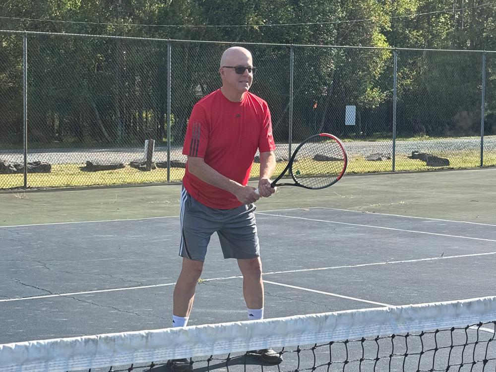 Stephen McNamara holds a tennis racket and prepares to return a shot on an outdoor tennis court, with a fence visible in the background.