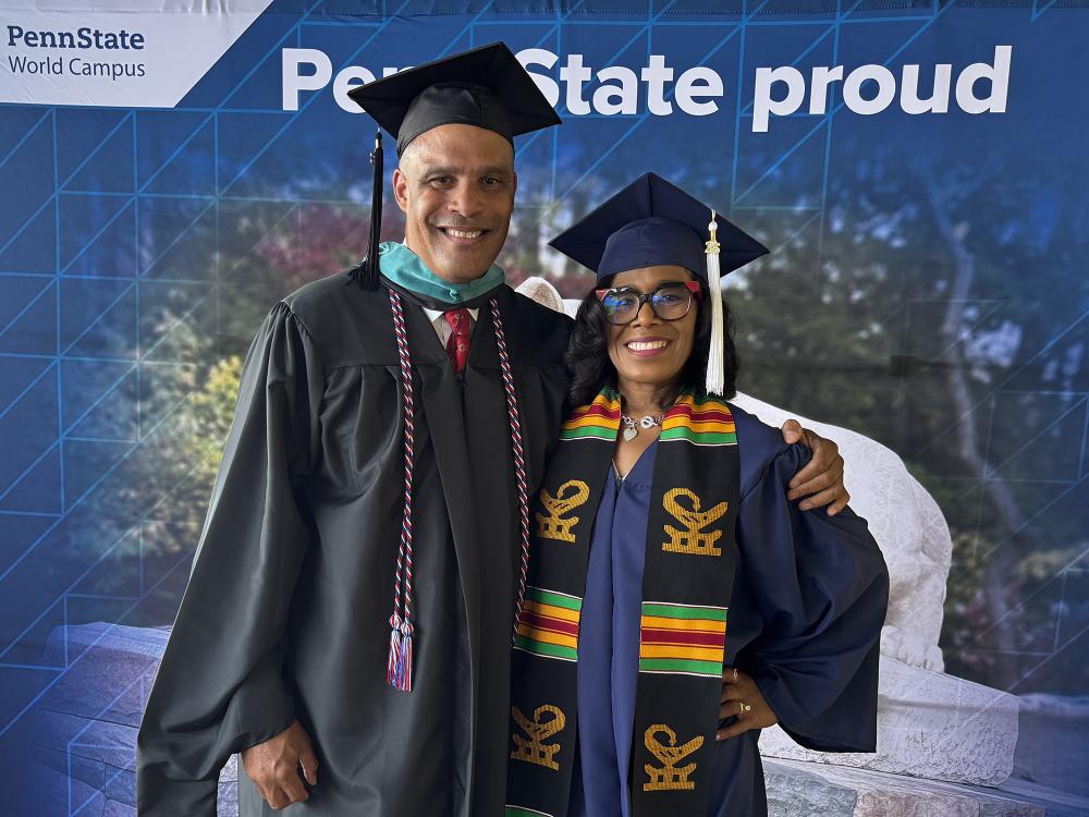 A man and woman wearing graduation regalia pose in front of a Penn State photo background.