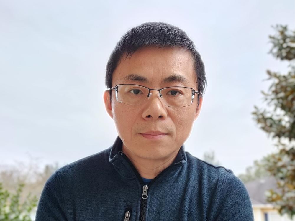An asian man with glasses and short dark hair wearing a dark blue zip front shirt