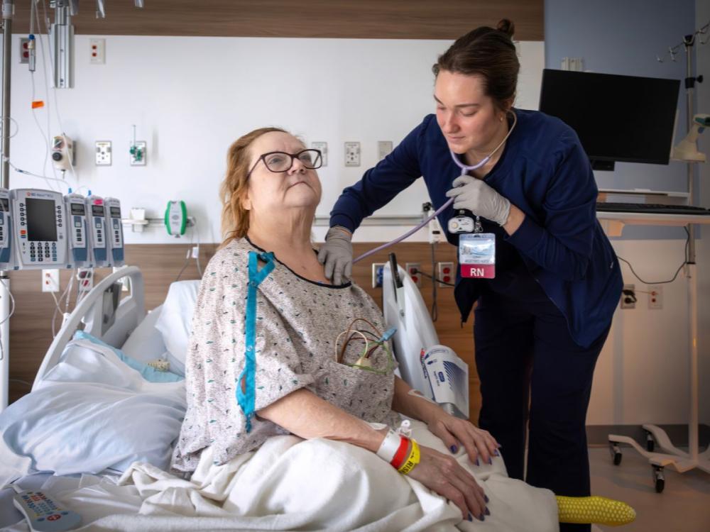 A nurse examines a hospitalized patient using a stethoscope while the patient sits upright in a hospital bed.