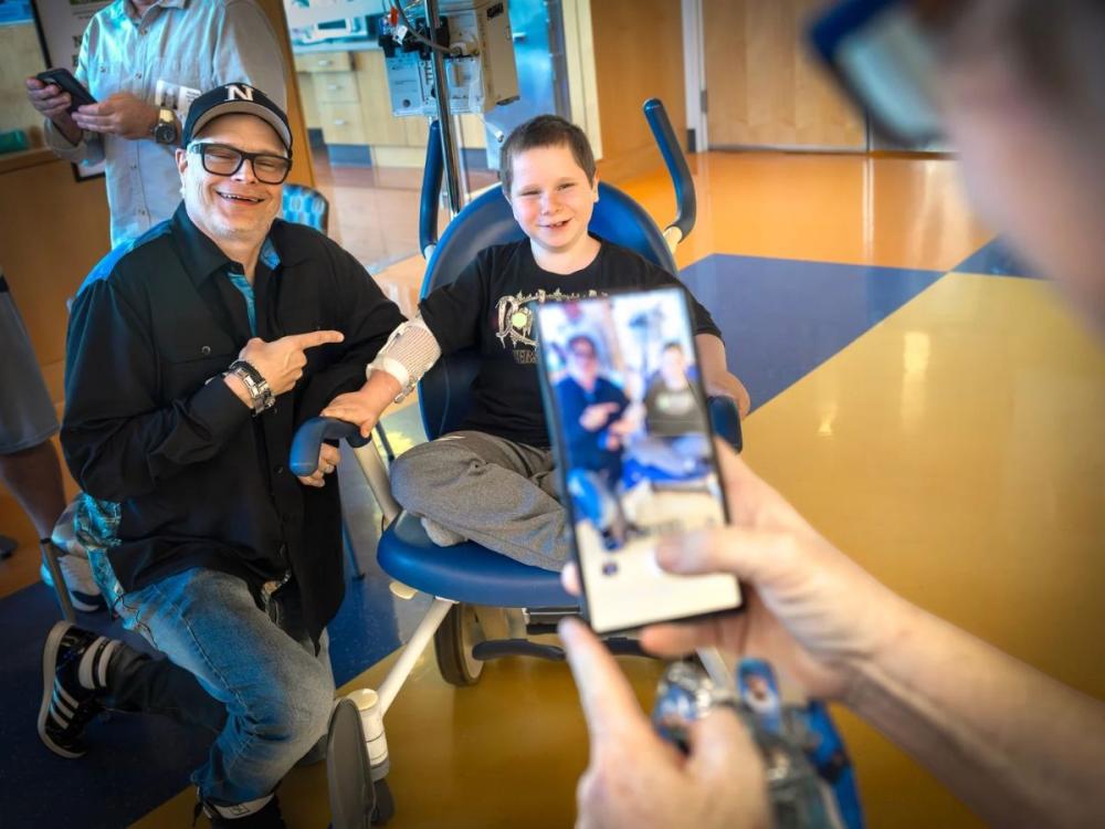A man and a smiling young boy sit together in a hospital treatment area, with the man pointing toward the boy’s bandaged arm as another person photographs them with a smartphone.