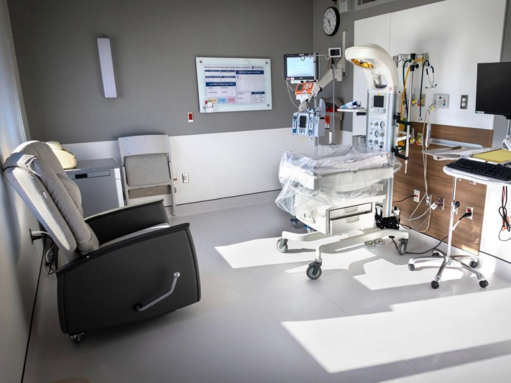 A private neonatal intensive care unit (NICU) room at Penn State Health Lancaster Medical Center featuring an infant radiant warmer with monitoring equipment, IV pumps, and respiratory support equipment beside a computer workstation. A cushioned recliner and chair provide space for parents, while large windows fill the room with natural light.