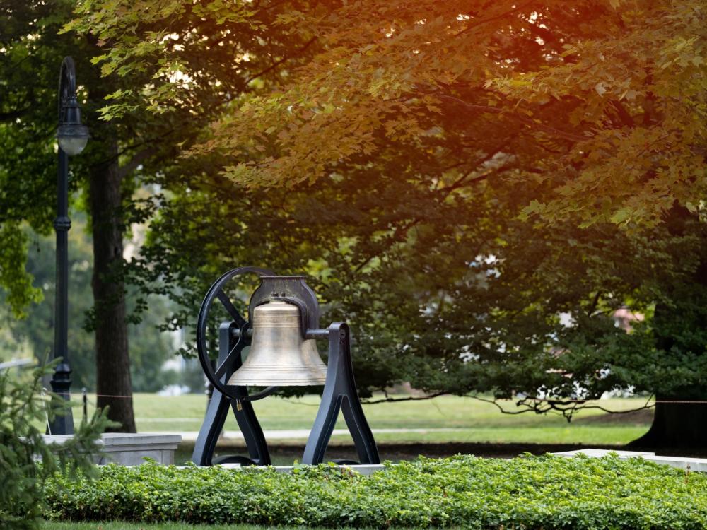 A picture of a large bell under a tree with sun shining through autumn leaves